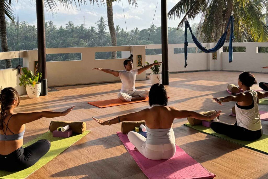 A group of people practice yoga on colorful mats in a sunlit, open-air studio with wooden floors and palm tree views. The instructor leads the class in a seated pose with arms extended, while participants follow along using props like bolsters and blocks.