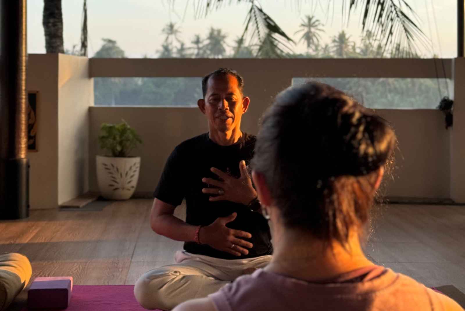 A yoga instructor sits cross-legged on a wooden floor, demonstrating a breathing or meditation technique with hands placed over his chest and abdomen, while a student observes from behind. The setting is a sunlit studio with large windows overlooking palm trees, and yoga props like a block and rolled mat are visible nearby.